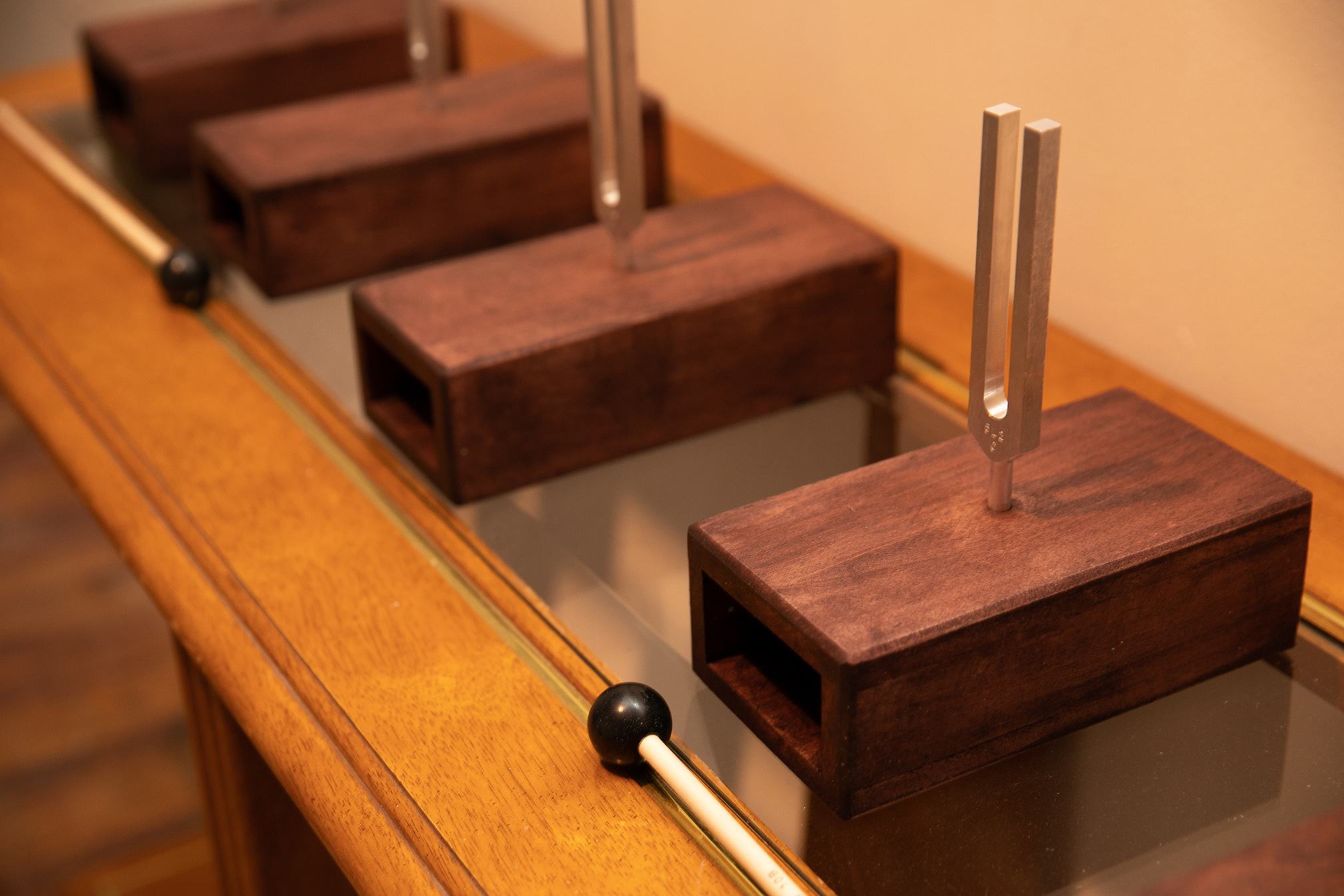 Extreme detail of a small wooden table with five tuning forks set in resanator boxes and two mallets set up on it against a white wall